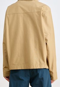 Beżowa lekka kurtka o luźnym kroju, z szwem na tyle i długimi rękawami, zestawiona z ciemnoturkusowymi spodniami.