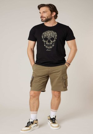 Man met een zwart T-shirt met een bloemige schedelprint, kaki cargoshorts, witte sokken en beige hoge sneakers, staand met handen in de zakken.
