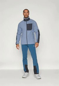 Giacca in pile di colore azzurro chiaro con dettagli blu navy, dotata di colletto alto, zip integrale e tasca sul petto. Indossata con pantaloni blu e scarpe bianche.
