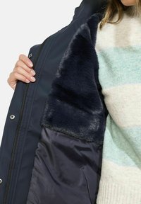 Manteau bleu marine présentant une doublure intérieure en fausse fourrure. Fermeture éclair avec boutons-pression, affiché partiellement ouvert révélant une texture douce.
