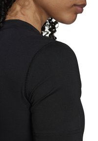 Gros plan sur l'épaule et le cou d'une personne portant un t-shirt noir ajusté à manches courtes avec des coutures visibles et des cheveux bouclés.