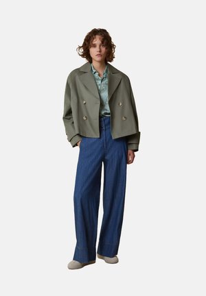 Giacca corta verde oliva con bottoni doppi, abbinata a una camicia a fantasia azzurro chiaro e a pantaloni in denim blu larghi.