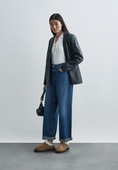 Μαύρο δερμάτινο σακάκι πάνω από ένα λευκό ριμπάριστο μακρύ μπλουζάκι, σε συνδυασμό με μπλε τζιν ψηλής μέσης και φαρδιάς γραμμής και καφέ χνουδωτά παπούτσια χωρίς κορδόνια. Συμπεριλαμβάνεται μαύρη τσάντα.