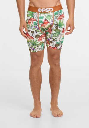 Mann, der bunte Boxershorts mit einem Muster aus Waldpilzen und Blättern trägt, steht barfuß vor einem weißen Hintergrund.