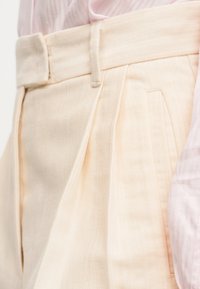 Μπεζ πλισέ παντελόνια με υφή υφάσματος, διαθέτοντας ίσια μέση και πλαϊνές τσέπες για μια δομημένη εφαρμογή.