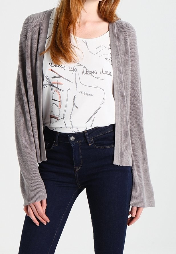 Hellgraue, offen gestrickte Strickjacke über einem weißen T-Shirt mit abstrakten Linien, kombiniert mit dunkelblauen, eng anliegenden Jeans. Lange Ärmel und lockere Passform.