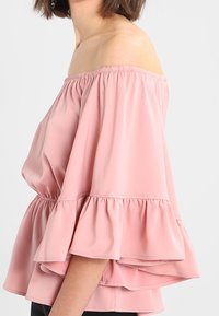 Top dénudé d'épaule en tissu rose doux, avec des manches évasées et un ourlet volanté, offrant une texture douce et légère et un col froncé.