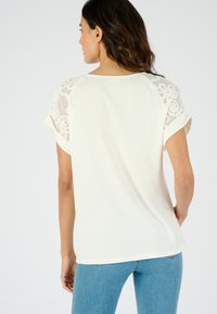 Top color crema con mangas cortas de encaje bordado y puños enrollados, con un corte holgado y un dobladillo trasero sutil. Lleva con jeans azules.