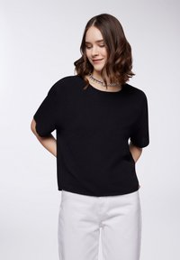 T-shirt nera corta con maniche larghe, collo rotondo, tessuto texturizzato, abbinata a pantaloni bianchi, con un design semplice senza motivi.