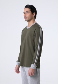 Camicia a maniche lunghe verde oliva con collo rotondo e polsini a coste grigi; presenta due righe bianche lungo ogni manica. Indossata con pantaloni grigi.