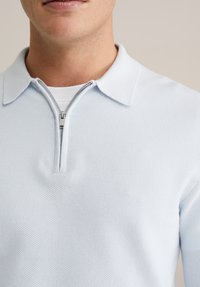 Svetlomodri pulover polo iz teksturirane tkanine, z ovratnikom na polovični zadrgo in kratkimi rokavi. Gladek, zračen dizajn, primeren za plastično oblačenje.