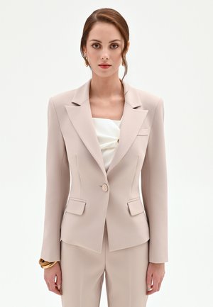 Donna che indossa una giacca beige aderente e pantaloni abbinati, con una maglietta bianca sotto e braccialetti d'oro al polso.