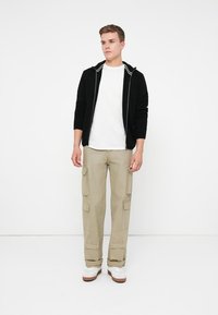 Musta vetoketjuhuppari, valkoinen t-paita, khaki cargo-housut, joissa on useita taskuja, valkoiset lenkkarit ruskeilla pohjilla, seisoo yksinkertaisella taustalla.