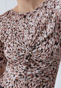 Primer plano de una persona que lleva un vestido estampado con un detalle de nudo retorcido en el pecho en tonos de marrón, negro y blanco.