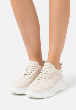Pieds chaussés de baskets en cuir beige avec des semelles blanches épaisses, lacées, affichant la marque "Copenhagen" sur les languettes et les côtés.