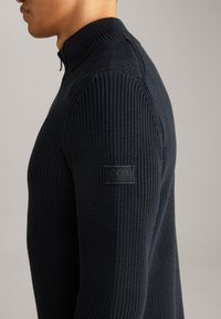 Strickpullover mit hohem Kragen in dunkelmarine Blau und mit strukturiertem Muster. Verfügt über einen Reißverschluss und ein kleines schwarzes Etikett am linken Ärmel.