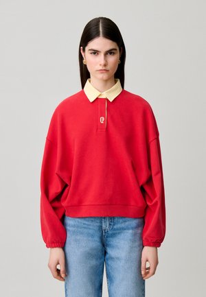 Mujer con cabello lacio y oscuro lleva un suéter rojo de manga larga con cuello beige y vaqueros azul claro, de pie frente a un fondo liso.