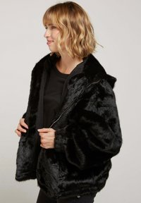 Veste en fausse fourrure noire avec capuche, dotée d'une fermeture éclair, de larges manches et d'une texture douce. Portée sur une chemise noire.