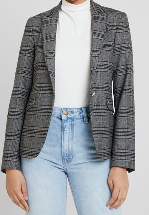 Persoon draagt een grijze geruite blazer over een witte coltrui en lichtblauwe hoge taille jeans, staande tegen een effen achtergrond.