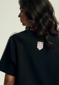 Czarna, oversize koszulka z różowymi paskami na ramionach, z białą i różową naszywką "COLLEGE 89" z gwiazdkami na plecach. Gładka tekstura materiału.