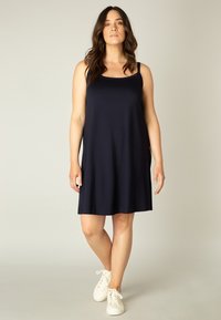 Marineblauwe mouwloze jurk van een zachte, vloeiende stof met een ontspannen pasvorm, voorzien van dunne schouderbandjes en een knielange zoom. Draagt men met witte sneakers.