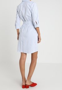 Abito camicia a righe blu chiaro con vita con cintura, maniche a 3/4 e colletto abbottonato. Indossato con scarpe rosse slip-on.