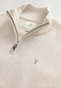 Beige half-zip sweater with ribbed collar, silver zipper, embroidered deer logo on chest, and Next brand label inside collar.
