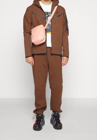 Brauner Zip-Hoodie mit schwarzen Akzenten, passende braune Hose, weißes T-Shirt und rosa Umhängetasche. Schwarze und rote Sneaker.