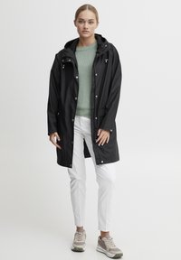 Svart vattentät regnjacka med huva, tryckknappar, sidofickor; matchad med en ljusgrön stickad tröja, vita byxor och grå sneakers.