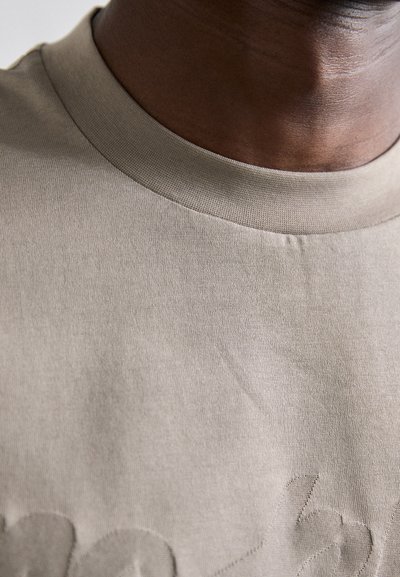 Beige t-skjorte med ribbet rund hals, med teksturert broderi på forsiden. Glatt stoff, minimalistisk design, nærbilde av halslinning og bryst.