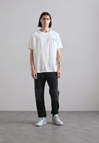 Hvit bomull t-skjorte med et lite svart trykt grafisk motiv, sammen med svarte bukser med teksturerte paneler. Grå sneakers fullfører antrekket.