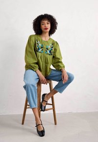 Zelená vyšívaná blůza s nabíranými rukávy, kombinovaná s světle modrými džíny a černými lakovanými Mary Jane botami, sedící na dřevěném stoličce.