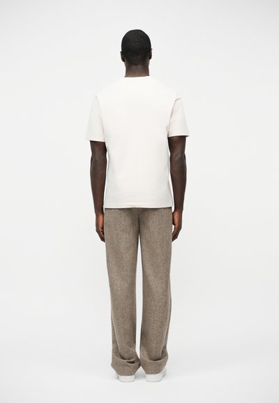 Άνδρας που στέκεται με την πλάτη προς το μέρος μας, φοράει απλό λευκό κοντομάνικο πουκάμισο, χαλαρό μπεζ παντελόνι και λευκά αθλητικά παπούτσια, σε λευκό φόντο.