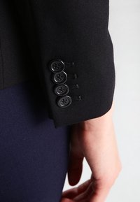 Manga de blazer de tela negra con cuatro botones negros y ojales estrechos; textura suave y ajuste estructurado.