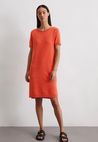 Robe à manches courtes en tissu tricoté orange vif, coupe décontractée, longueur genou, avec un col rond et un ourlet côtelé. Associée à des sandales noires.