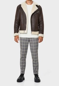 Brown leather jacket with shearling lining, zippered closure, and two side pockets, paired with gray plaid tapered trousers and black shoes.
