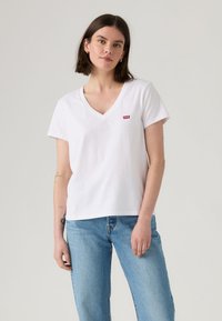 T-shirt de algodão branco com decote em V e mangas curtas. Apresenta um pequeno logótipo vermelho da Levi's no peito esquerdo. Usado com jeans azul claro.