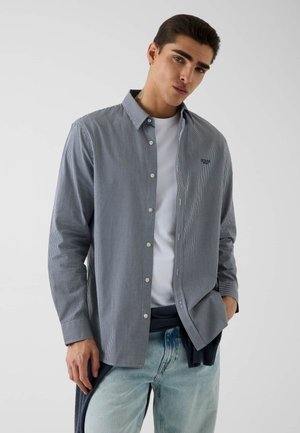 Jeune homme portant une petite chemise à carreaux boutonnée sur un t-shirt blanc et un jean bleu clair, debout avec une main dans la poche.