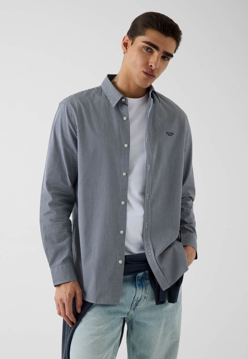 Jeune homme portant une petite chemise à carreaux boutonnée sur un t-shirt blanc et un jean bleu clair, debout avec une main dans la poche.