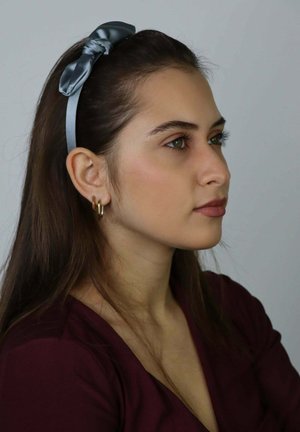 Grey satin headband with a bow detail, smooth texture. Model wears gold hoop earrings and a deep burgundy top, facing slightly to the side.