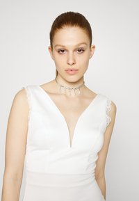 Vestido branco com um decote em V profundo, com detalhes em renda nos ombros e uma silhueta ajustada. Acompanhado de um colar de pérolas com pendentes.