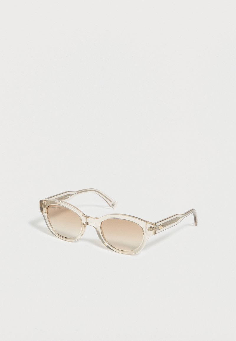 Lunettes de soleil à monture beige transparente avec des verres dégradés marron, inclinées sur une surface blanche.