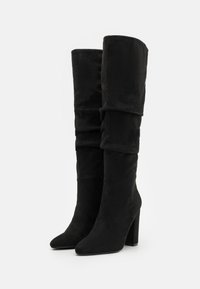 Botas negras por encima de la rodilla hechas de suave ante, con un diseño arrugado, punta afilada y tacón alto grueso. Textura suave sin adornos.