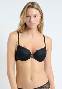 Sujetador negro con aros, detalles de encaje, bordes en picos y tirantes ajustables. Cuenta con un pequeño elemento decorativo en el centro delantero.