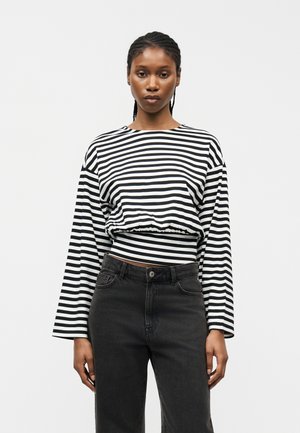 Donna con capelli intrecciati che indossa una maglia a maniche lunghe a righe orizzontali bianche e nere e jeans neri a vita alta, in piedi su uno sfondo bianco.