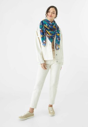 Foulard carré en tissu bleu arborant un motif coloré de véhicules. Le modèle porte une veste en denim blanche, un top crème et des baskets beiges.