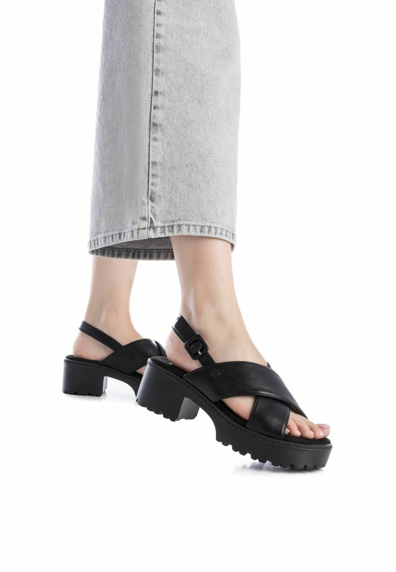 Sandalias de cuero negras con un diseño de tiras cruzadas, hebilla ajustable y tacones gruesos texturizados, combinadas con unos jeans de mezclilla gris claro.