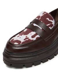 Debel, temno rjav usnjen loafer s črnim debelim podplatom in belim ter rjavim krznenim panelom na vrhu z vzorcem kravjega usnja.