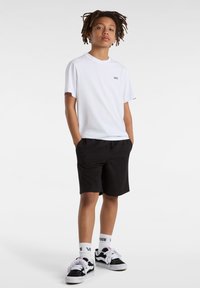 T-shirt bianca in cotone con logo nero, abbinata a pantaloni corti neri. Piedi in scarpe da ginnastica nere e bianche, in posizione sicura con le mani in tasca.