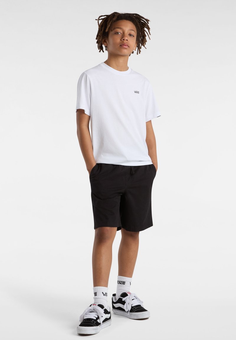 T-shirt bianca in cotone con logo nero, abbinata a pantaloni corti neri. Piedi in scarpe da ginnastica nere e bianche, in posizione sicura con le mani in tasca.